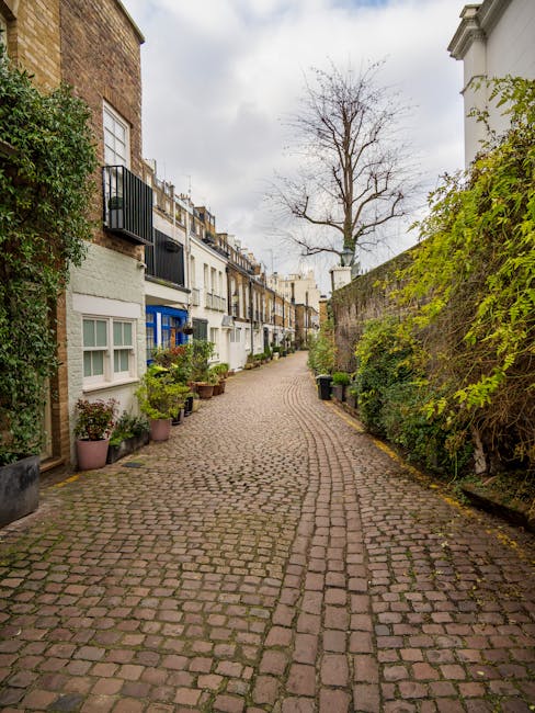 A narrow cobbled street in Fulham lined with terraced houses and small front gardens. On the left, potted plants and shrubs sit beneath white and pastel-colored facade windows, while on the right, a tall brick and stone wall is partly covered with green climbing plants and overhanging branches. The street curves gently ahead, with a leafless tree towering in the background under an overcast sky, suggesting early autumn or late winter. The scene captures a quaint, historic residential area that may present garden clearance challenges, emphasizing the need for careful outdoor maintenance and landscaping work, supported by Gardeners Fulham.